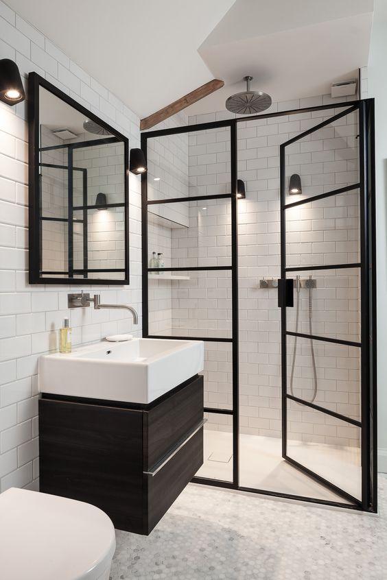 Banheiro com revestimento subway tiles branco, área do box com piso porcelanato branco e demais área com piso branco hexagonal em pequeno formato, box de vidro com molduras pretas, espelho com moldura preta e gabinete marrom escuro com cuba branca.