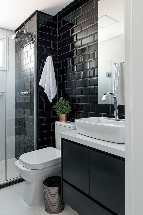Banheiro com azulejo subway tiles preto na parede principal e demais paredes pintadas de branco, louças brancas e armários com bancada branca e portas pretas.