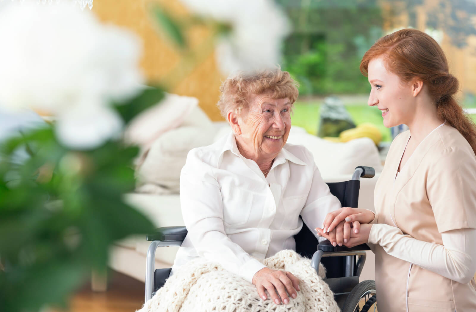 A senior woman in a senior living facility sitting in a wheelchair smiling and having a conversation with a nurse.