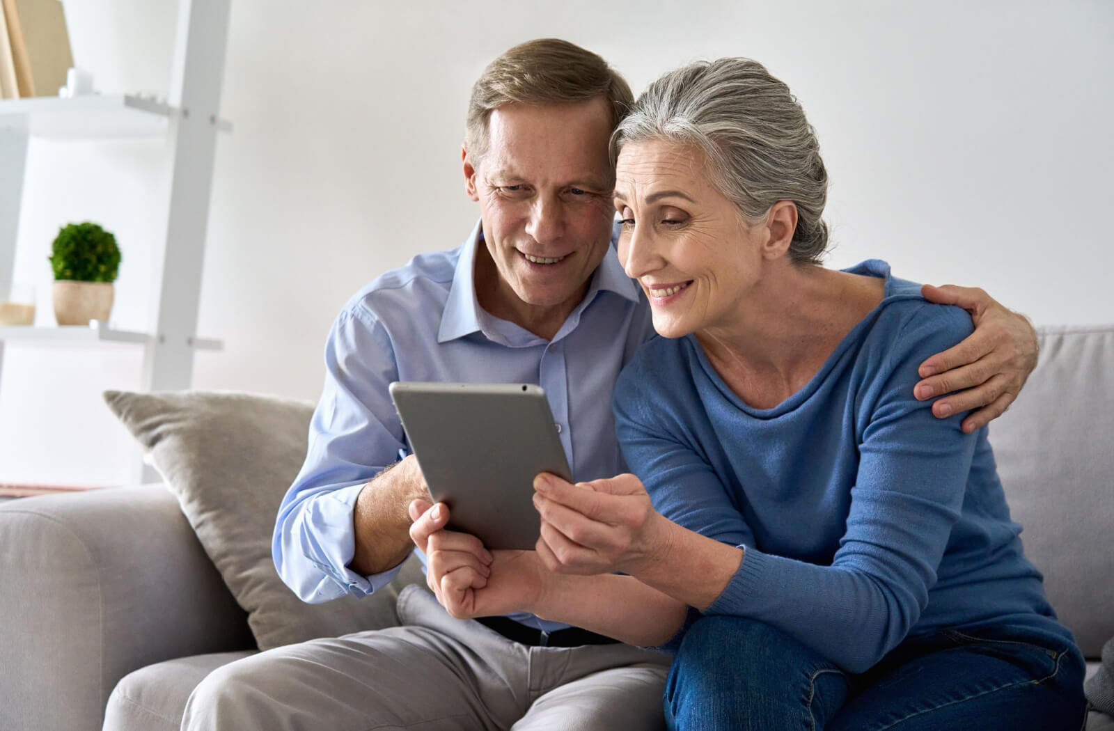 A man in a long polo shirt is sitting on a couch beside her mother in a blue shirt while he is showing a virtual tour of the services and amenities inside an assisted living facility.