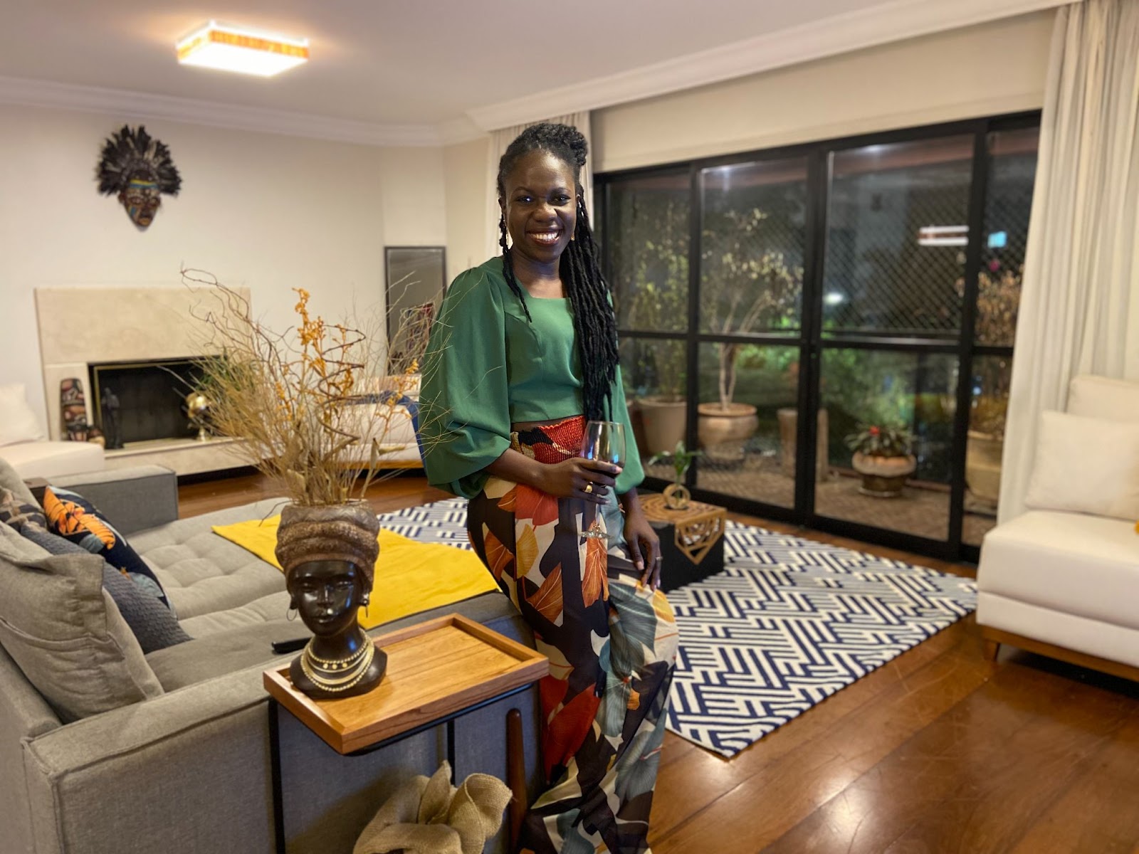 Imagem mostra Sauanne Bispo, uma mulher negra e jovem, sorridente. Ela está em uma sala ampla e espaçosa, com símbolos da cultura negra no ambiente e uma varanda cheia de plantas. 