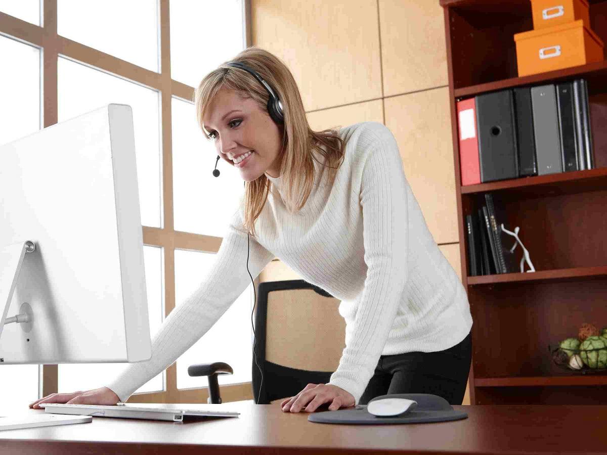 A woman dressed in formals wearing headphones, standing at a desk and working on a computer