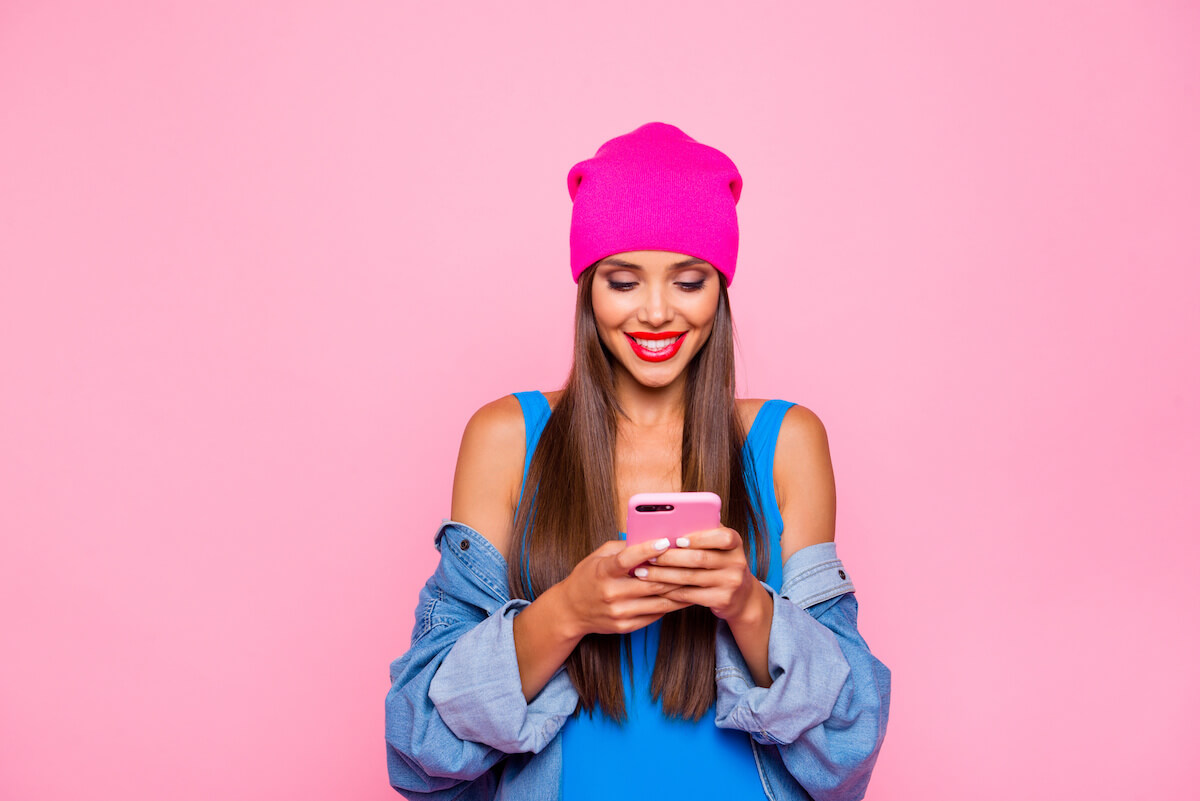 Woman in hot pink beanie types on phone in front of pink background