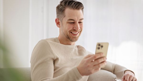 homem loiro com camisa bege sentado vendo celular e sorrindo