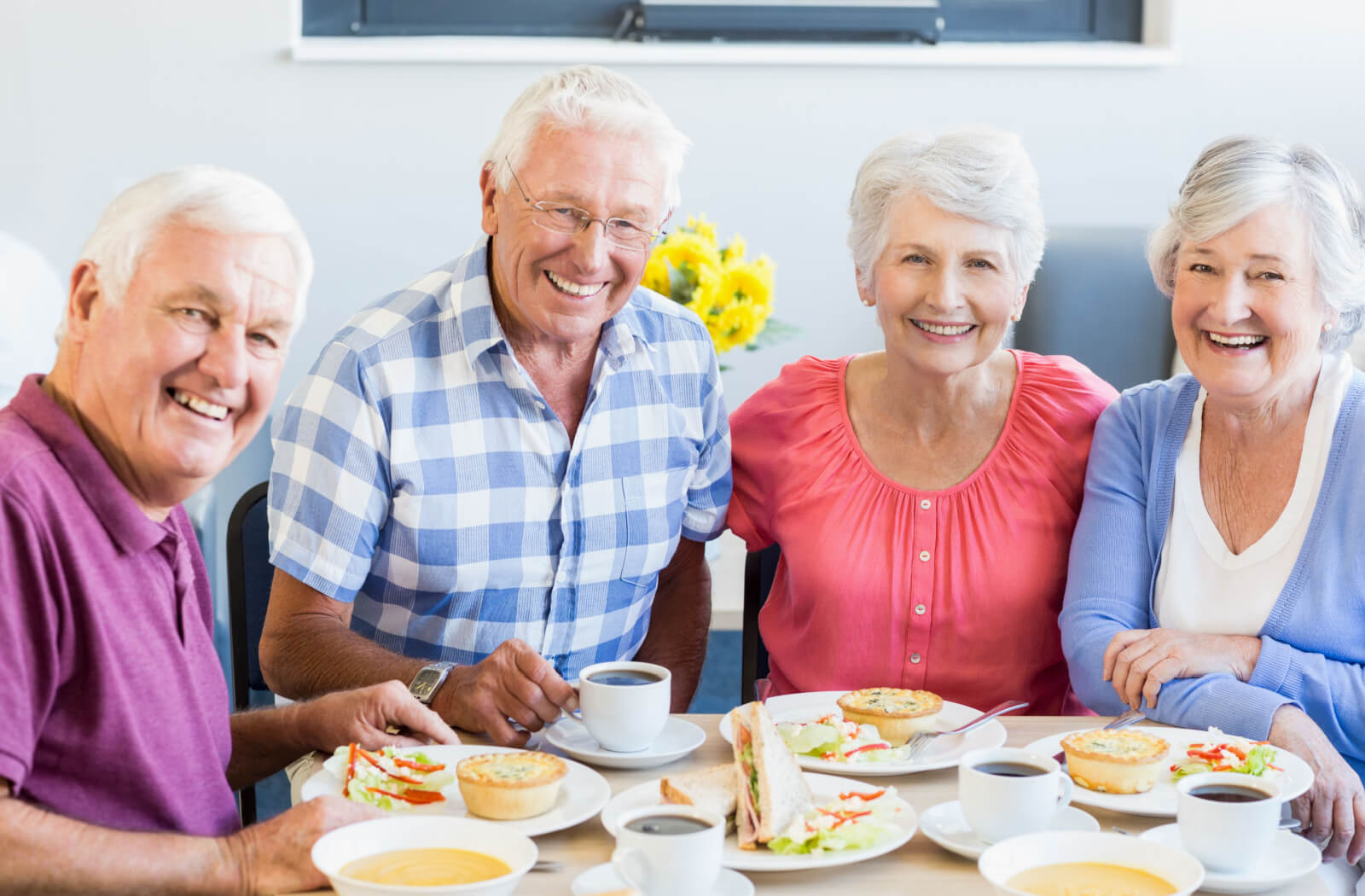 A group of seniors  sitting around a table, eating and having a sandwich and tea, while smiling  and looking directly at the camera.