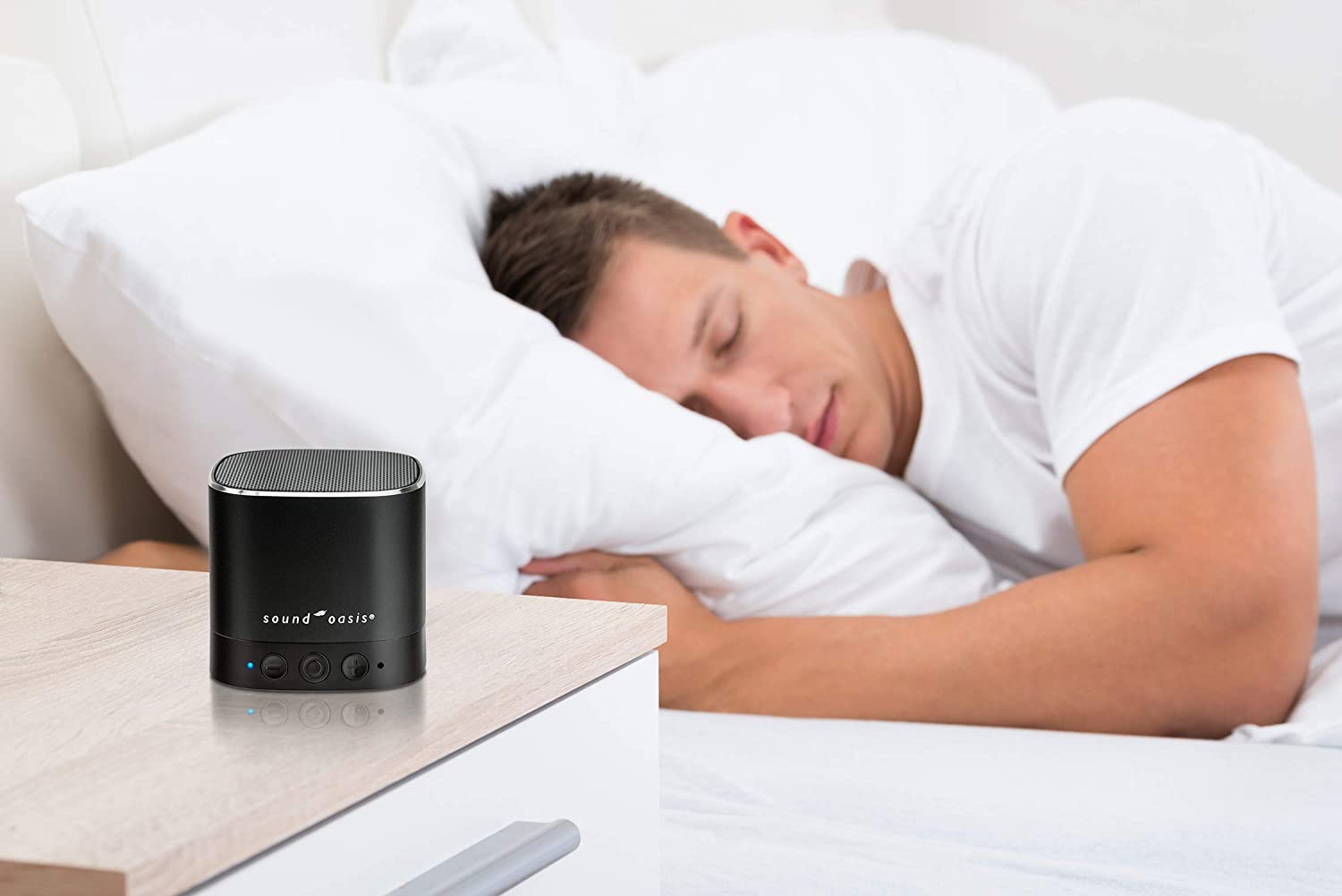 A Sound Oasis Bluetooth Sound Therapy System for tinnitus is on a bedside table. A man is sleeping in the background.