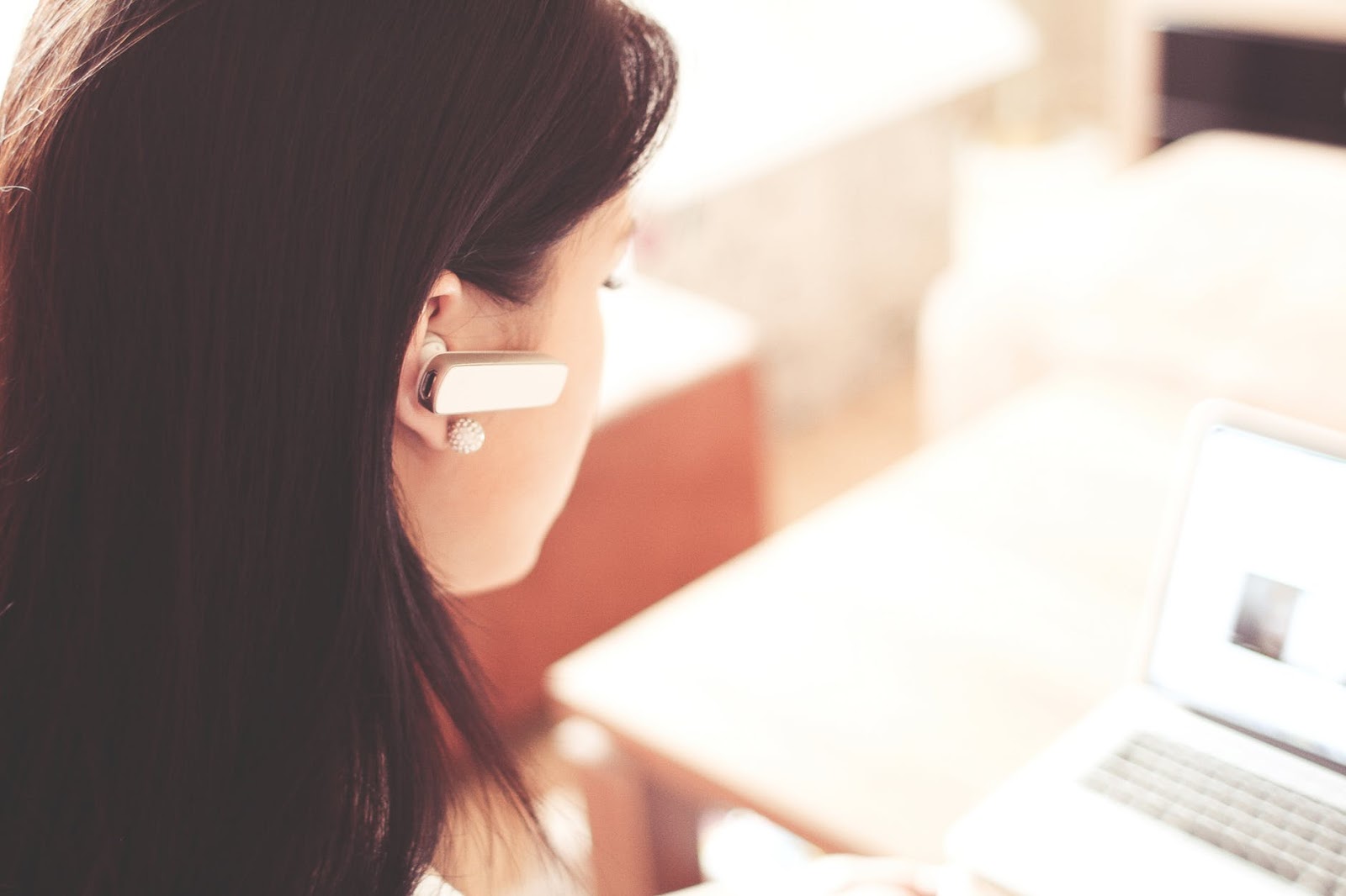 a woman facing her laptop with a wireless headset on her right ear.