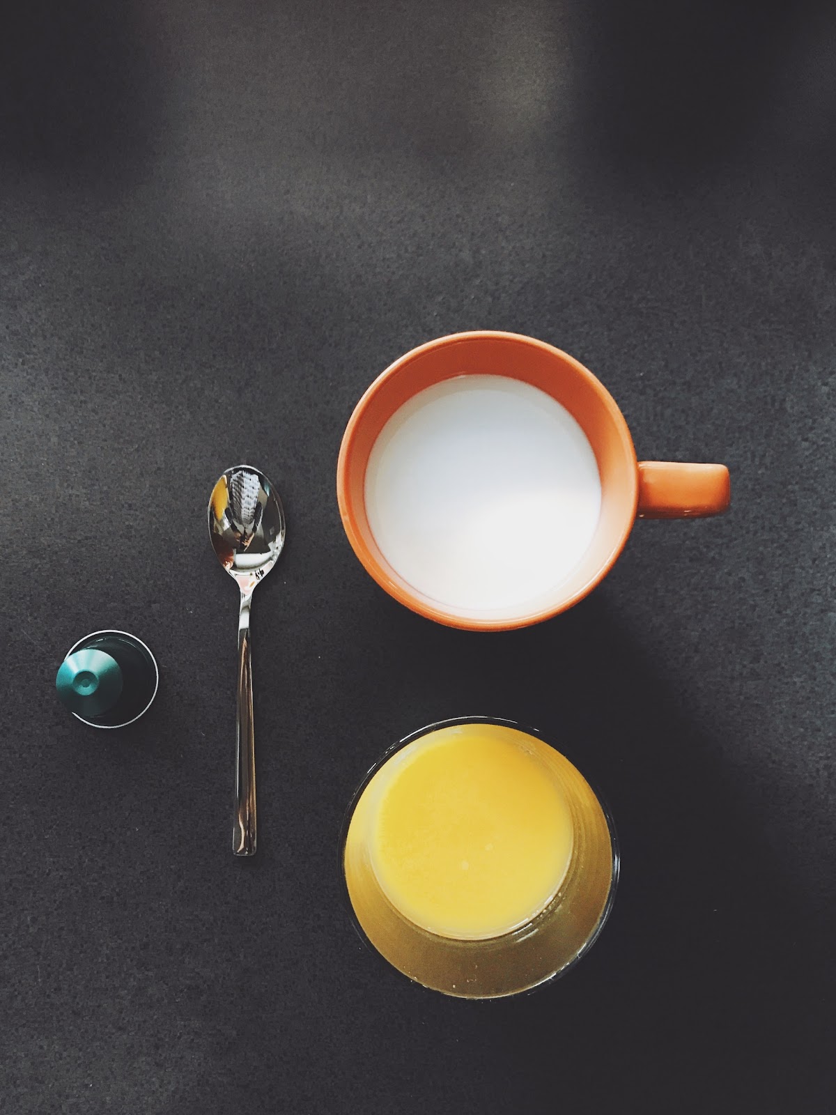 A glass of milk and a glass of juice sit on a table next to a spoon.