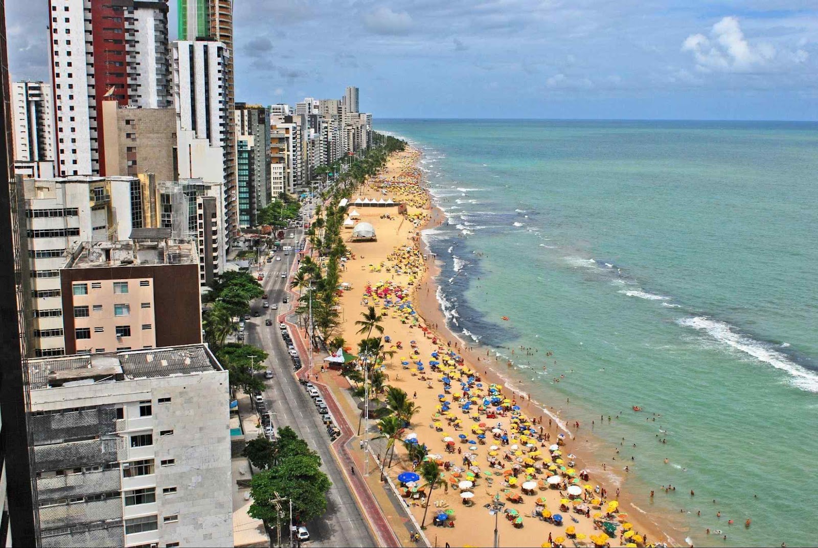 Orla da Praia da Viagem em Recife. A areia da praia está cheia de banhistas e os prédios da cidade contornam a orla.