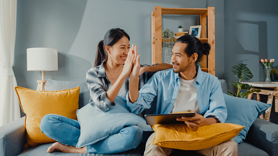 An Asian-American couple sitting on their couch in their living room, turning towards each other, giving each other high-five as they feel connected and strong in their relationships. Learn better communication skills in couple's counseling in Woodland Hills, CA. 91364