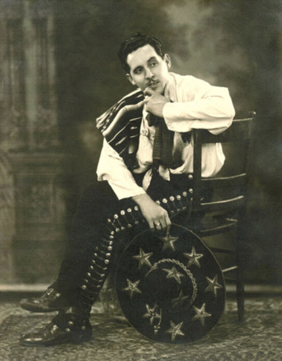 Black and white photo of a young Raoul Cortez sitting in a chair with a hand holding a sombrero and another hand touching his chin