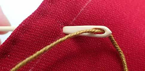 A wood bodkin is laced with the gold drawstring cords and is pilling one cord from the red side into the bag through the hole just made by the awl.