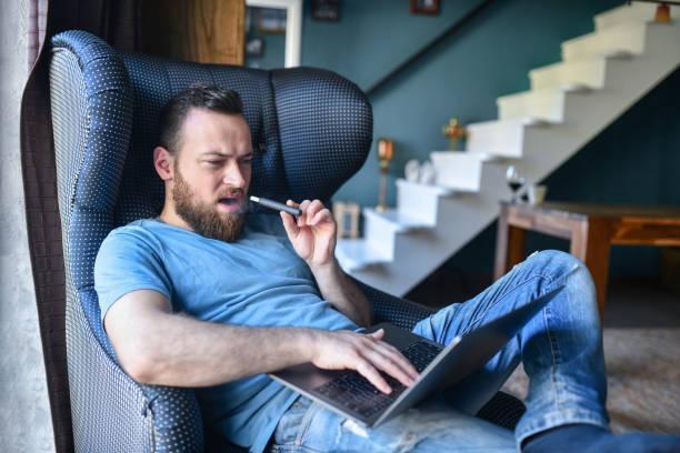 Bearded Male Freelancing From Home On Laptop And Smoking Electronic Cigarette Bearded Male Freelancing From Home On Laptop And Smoking Electronic Cigarette vaping at home stock pictures, royalty-free photos & images