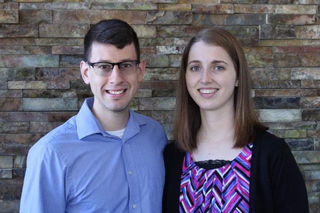 Two people standing in front of a brick wallDescription automatically generated