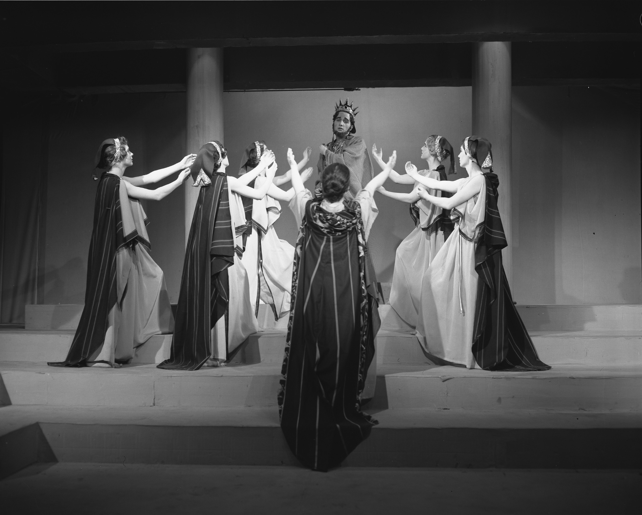 A circle of six Caucasian women stand on a marble staircase. They hold their arms up towards a king who stands at the top step. He has a black beard and a pointed golden crown. The women wear long flowing robes belted at the waist, and scarves over their hair.