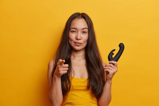 Mulher jovem confiante aponta o dedo indicador, segura o vibrador para estimular o clitóris com vibrações cintilantes, tem um dildo pessoal, isolado na parede amarela. brinquedo sexual para mulheres. Foto gratuita