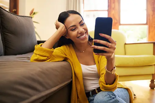 Woman Smiling At Her Phone Reading Twitter Selfie Captions While Sitting On The Floor Along a Sofa