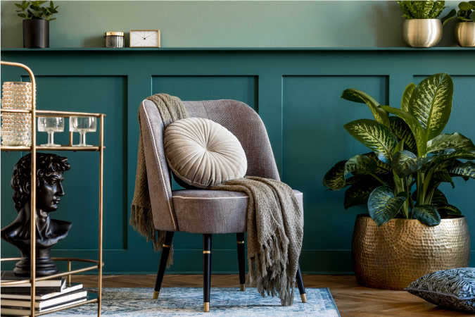 A close-up photo of a trendy living room. The wall is painted in two complementary blue-gray tones. There are plants arranged around the room in decorative metal pots. Stylish, retro furniture brings the room together.