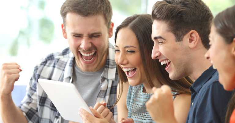 Emocionado grupo de cuatro amigos jugando al bingo online en línea desde una tablet en el interior de una casa