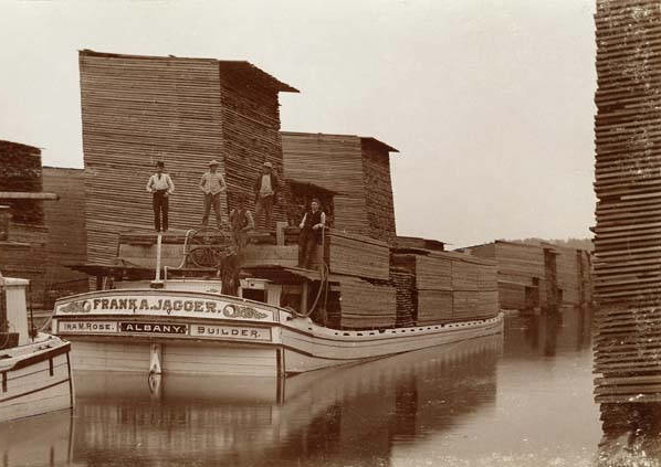 A flat boat with large, wooden boards piled on it floats in a narrow channel surrounded by more piles of wooden boards. A few men pose on the boat.