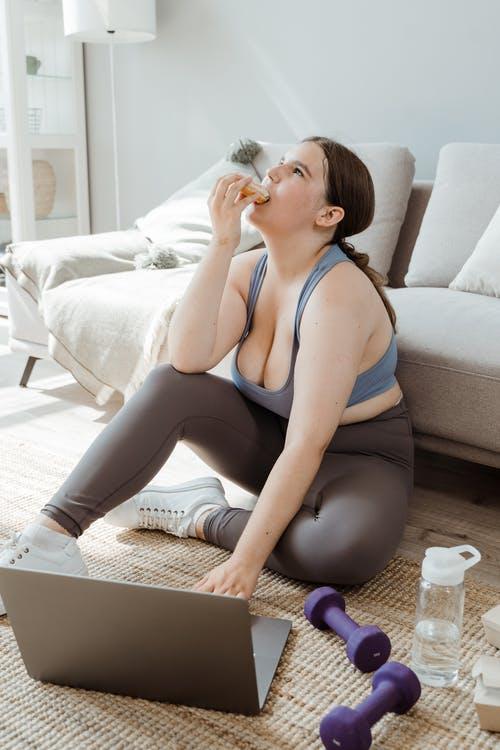 Woman in Blue Tank Top and Black Leggings Sitting on the Floor Besides Weights and Laptop and Eating Cake