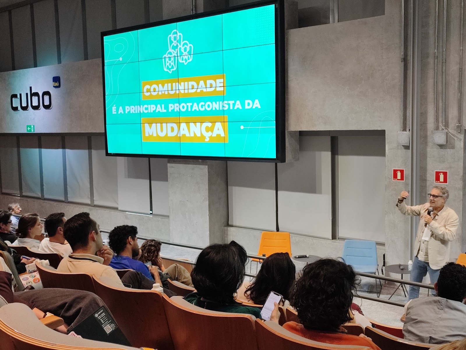 Foto da apresentação de Saulo Barreto, um homem branco, grisalho, de calça jeans e casaco da cor creme. Ele está em frente à plateia e de pé segurando um microfone, atrás dele está uma parede de concreto e mais acima um telão com uma apresentação de slides com o texto “Comunidade é a principal protagonista da mudança”.