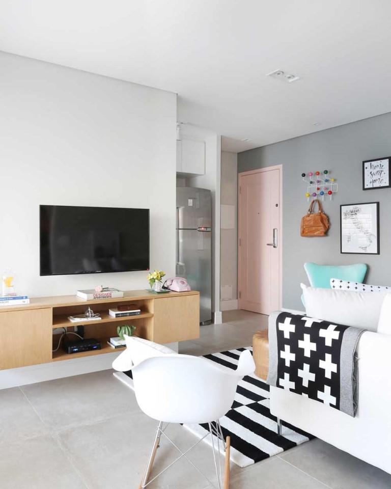 Sala com parede e piso cinza, porta rosa, rack amadeirada, poltronas branca e azul, tapete listrado branco e preto e sofá branco com almofadas coloridas.