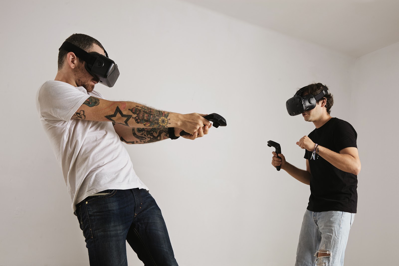 Foto de um pai jovem e um filho adolescente interagindo, em pé, e se divertindo com óculos de Realidade Virtual e consoles em mãos.