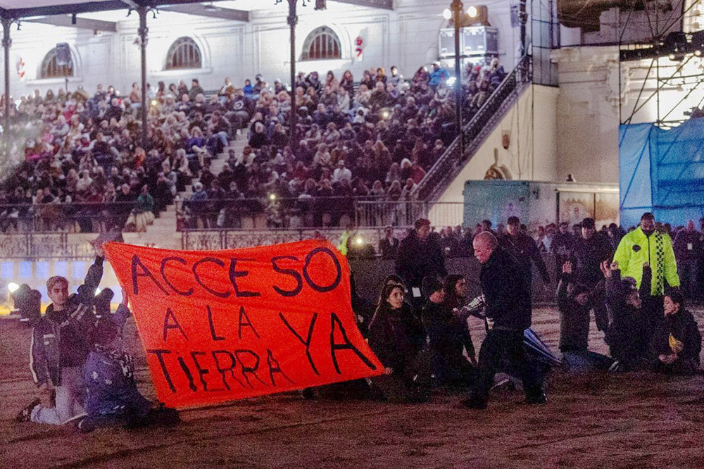 Protestos sit on the floor of a stadium with crowd in stands behind. They hold a red banner demanding Access to Land