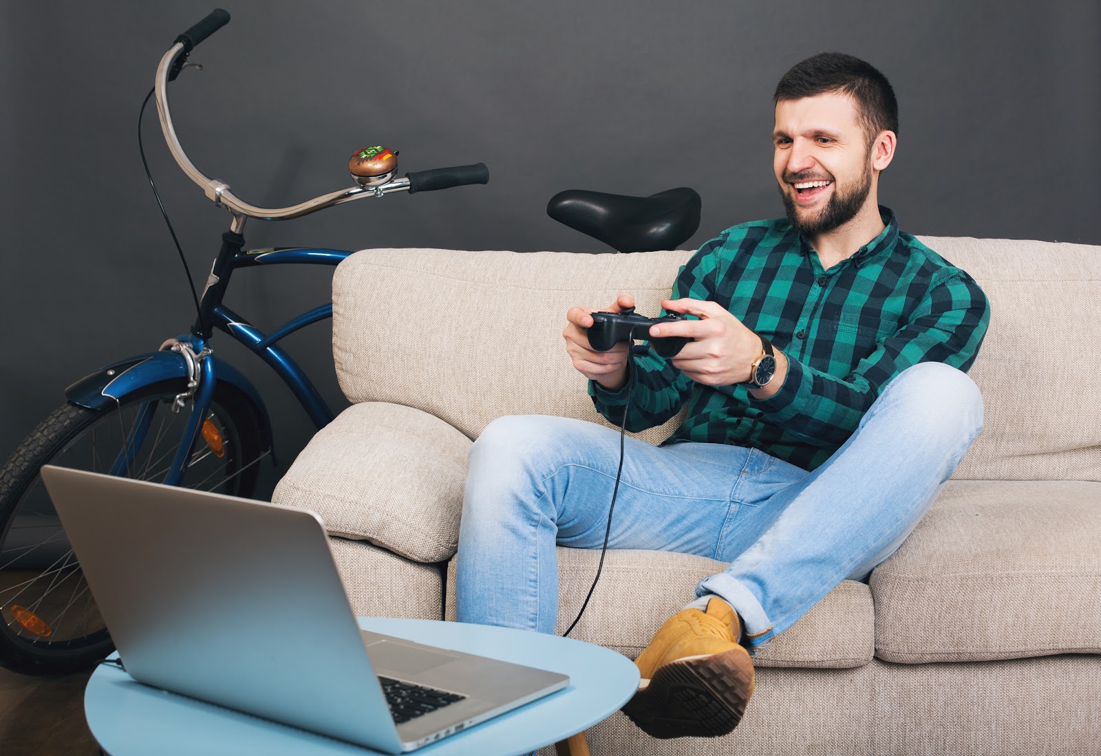 Foto de um homem jovem, com um console de videogame nas mãos, sentado em um sofá, de frente para um notebook e com uma bicicleta ao fundo, atrás do sofá. O objetivo da imagem é saber o que é SSD no notebook e sua importância para o bom desempenho gamer.