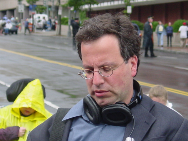 Man in rain jacket with glasses looks down with headphones around his neck people and police in rain gear are in the background. 