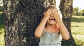 A child smiling behind a tree, covering her eyes playing hide and seek