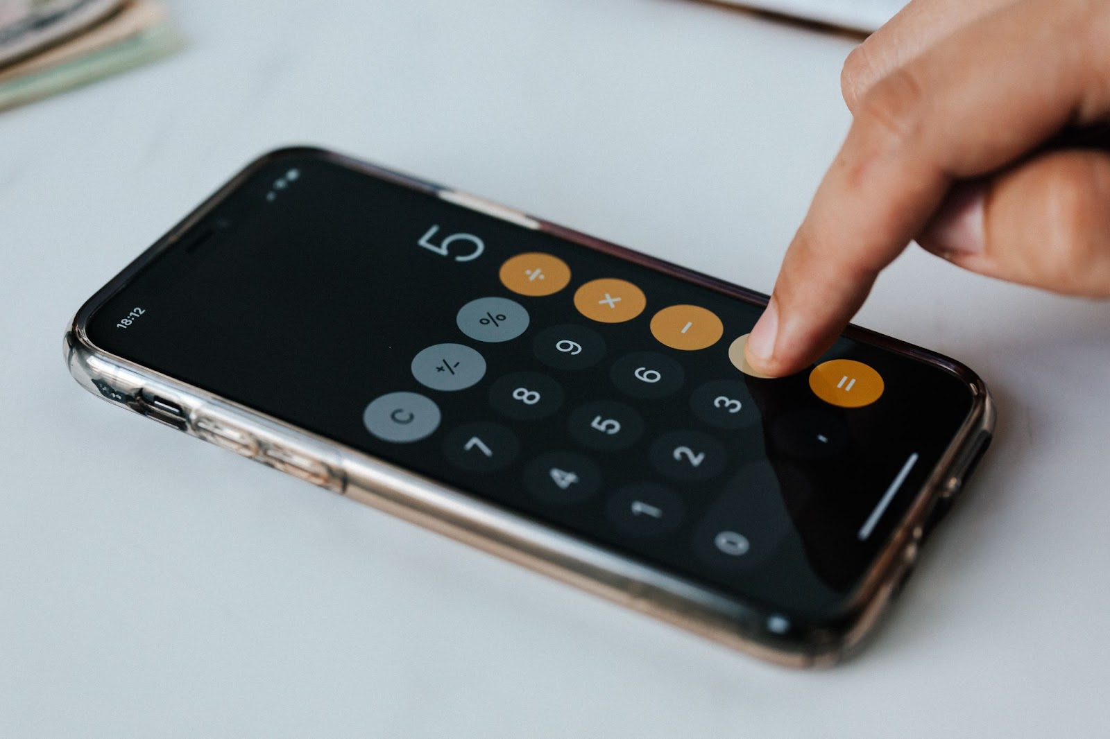 Na imagem, a mão direita de uma pessoa branca está digitando sobre um celular que mostra uma calculadora digital, com o número 5 sendo mostrado no visor. O celular está disposto sobre uma superfície clara, como uma mesa.