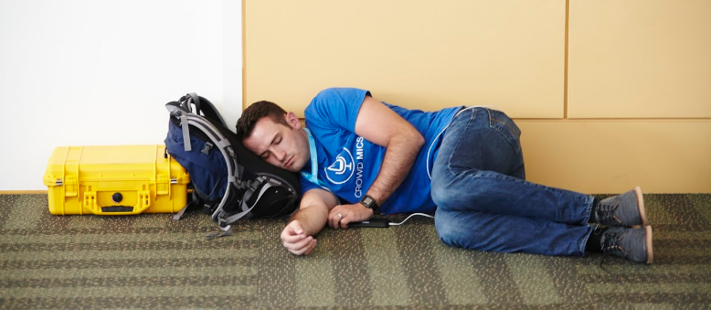 Person reclining against a wall, head resting on a backpack.