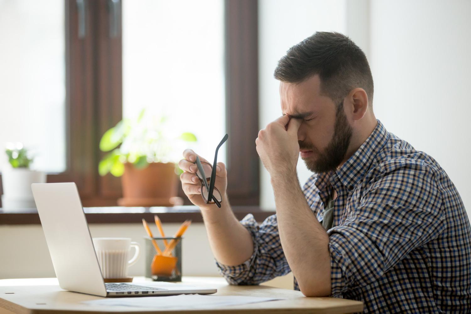 La imagen muestra a un hombre sentado frente a una mesa de madera. Está en el lado derecho de la imagen, con la mano derecha sostiene las gafas y con la izquierda presiona la zona entre la nariz y los ojos, como si estuviera muy cansado. Además, sobre la mesa es posible ver un cuaderno abierto, lo que nos da la impresión de que está trabajando.