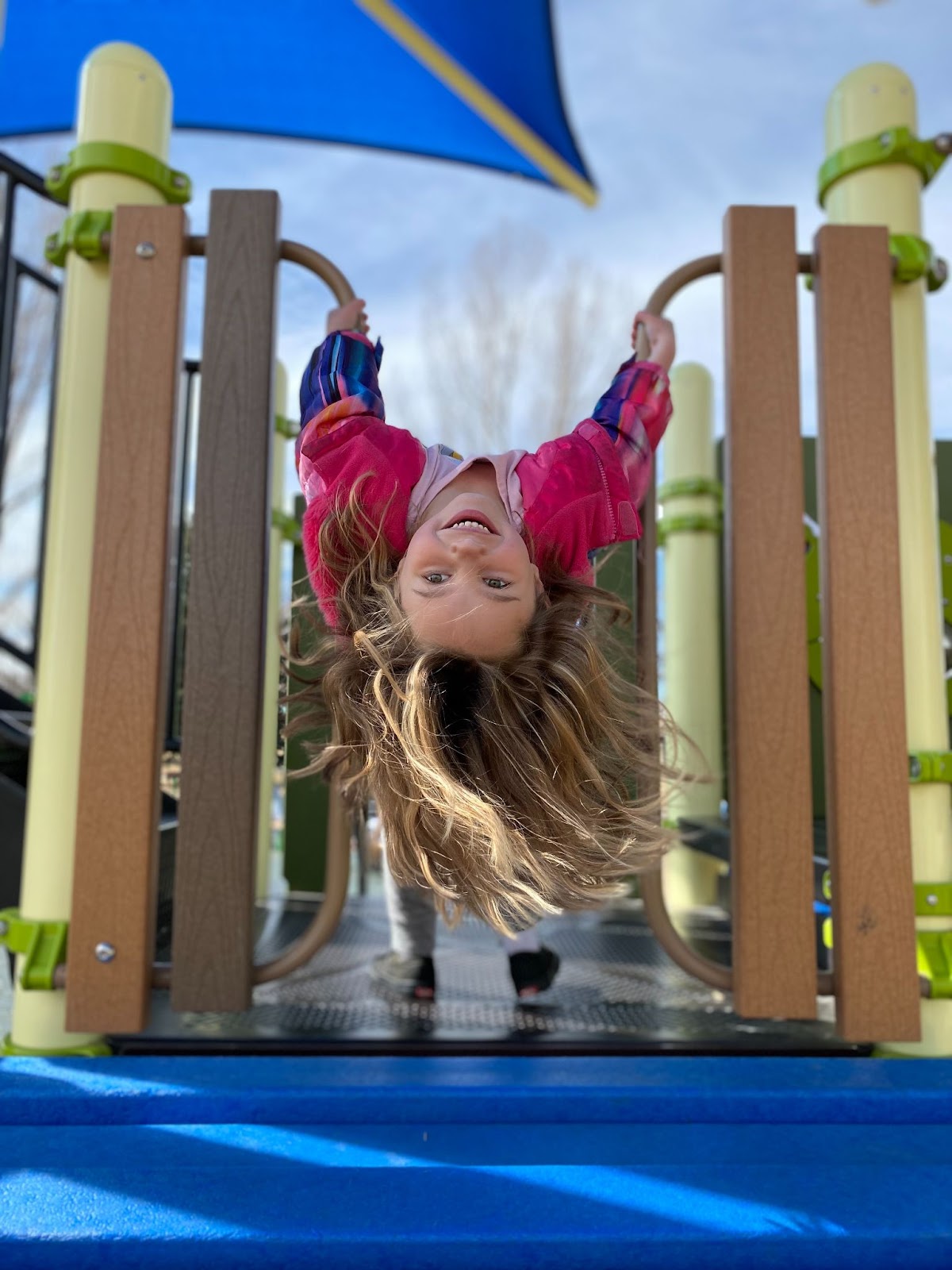 A little girl in a pink jacket hangs upside down on playground equipment