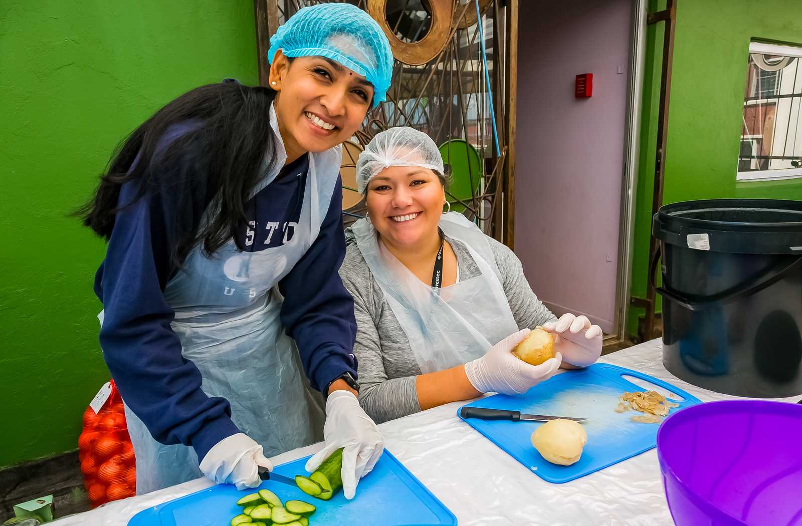 volunteers smiling while doing food prep on a bright green wall and purple bowl