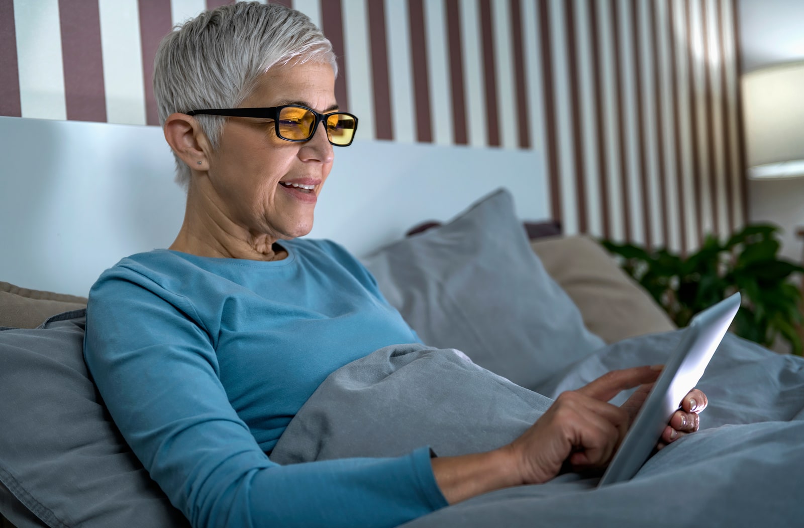 An older woman lying in bed wearing blue light blocking glasses while reading her tablet screen and touching it
