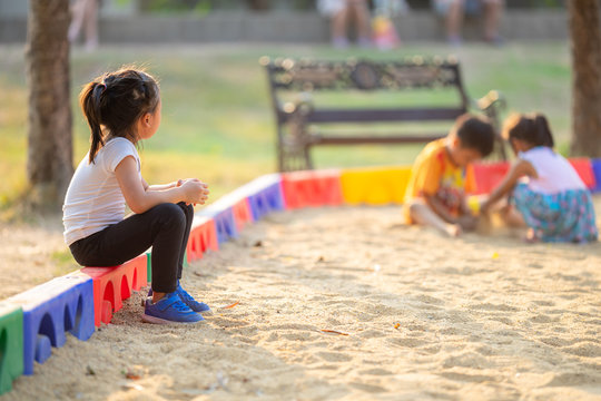 young girl sitting alone on the low wall of a playground sand area watching two other children in the distance play in the sand together.