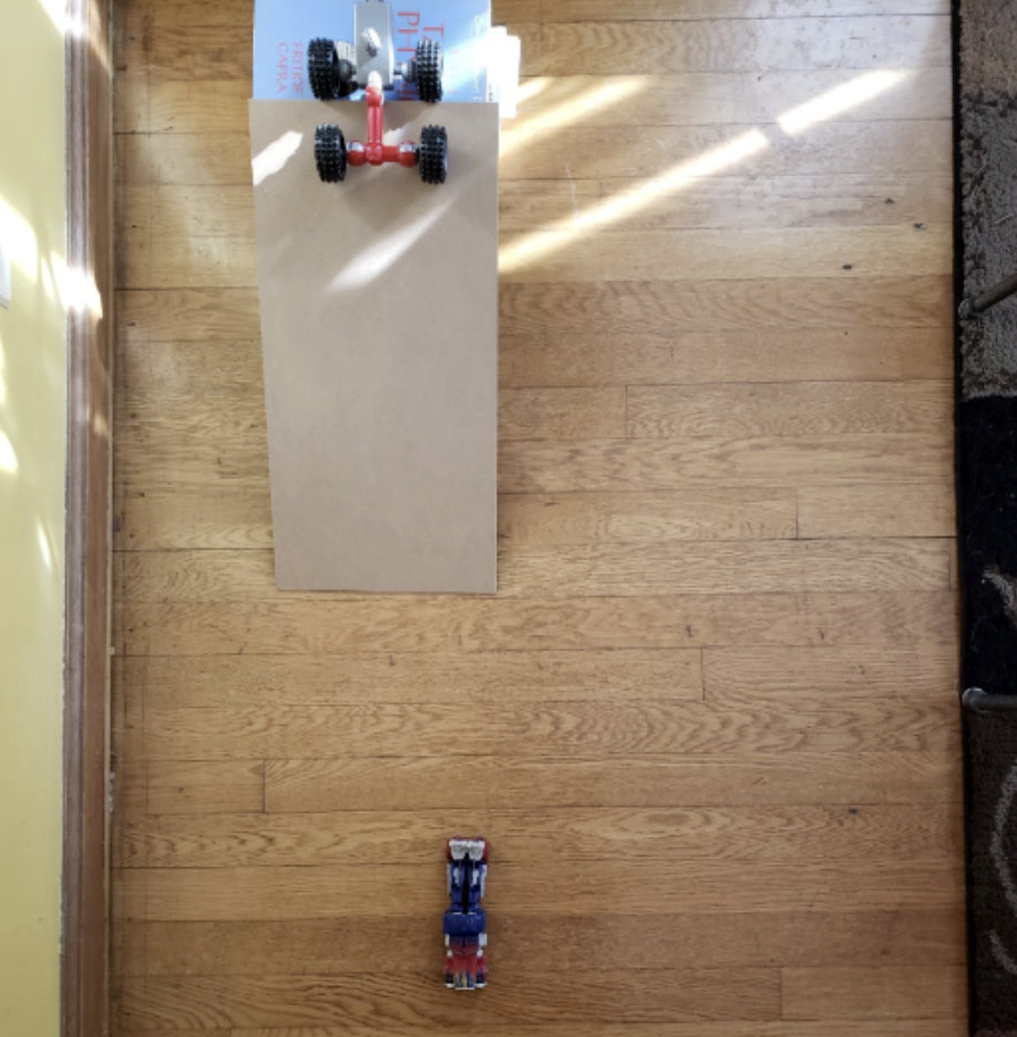 A toy car sits at the top of a ramp. Another toy car has already rolled down the ramp.