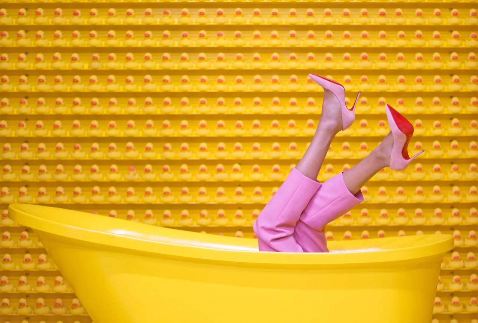 Una mujer vestida de rosa y con zapatos de tacón con suela roja, enseña sus pies metida en una bañera amarilla, sobre un fondo amarillo. Destazan así los zapatos Louboutin.