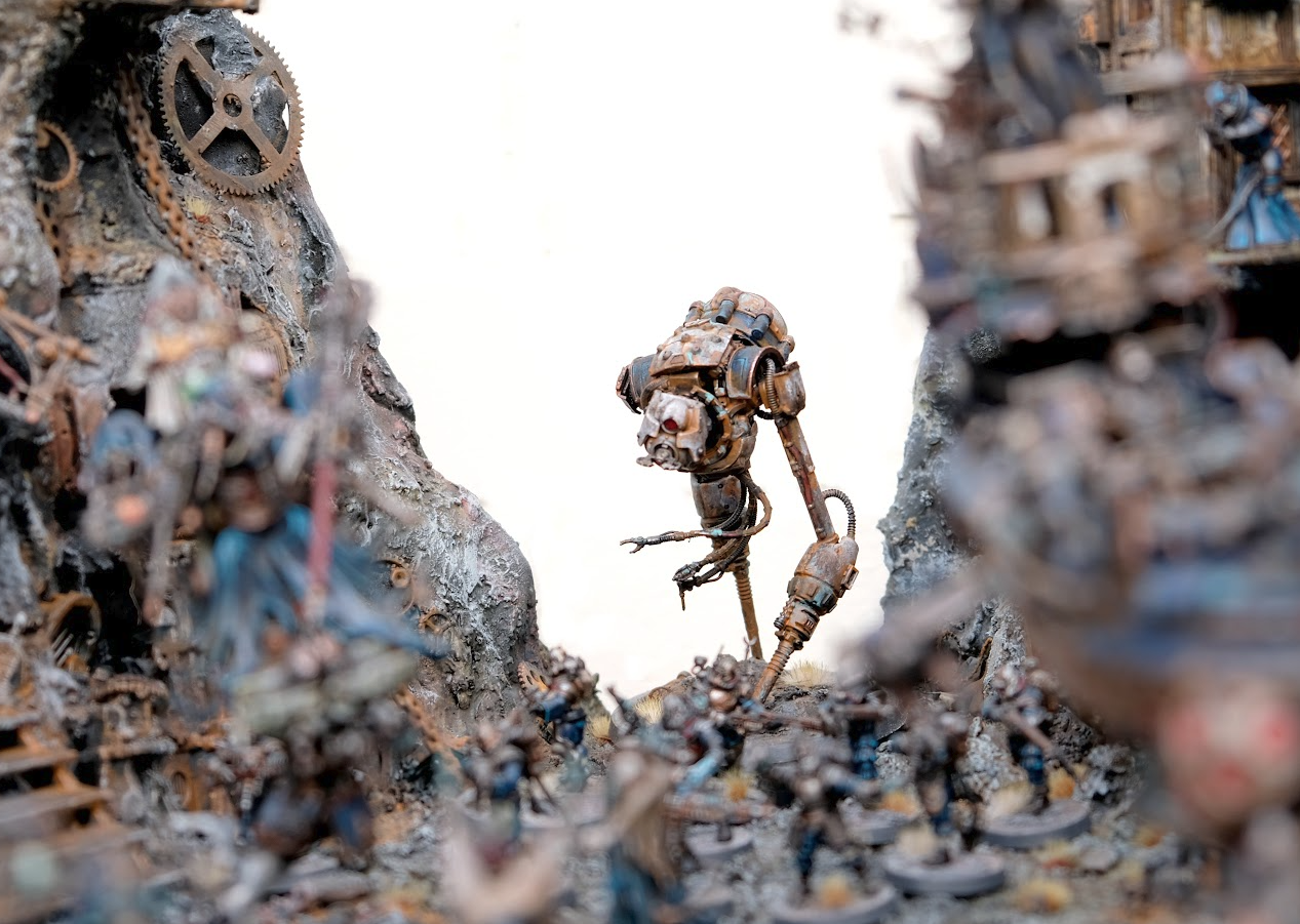 A tall robot on long striding legs is in focus at the back of an army diorama 