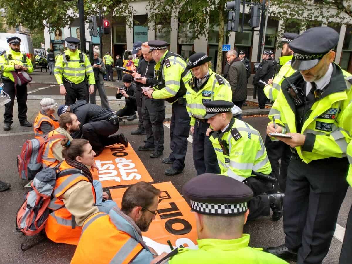 a line of police interview a line of activists knelt across a road to blockade it 