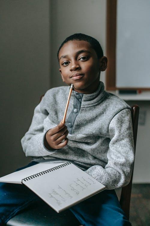 Focused African American boy sitting on chair and looking at camera while doing tasks in copybook
