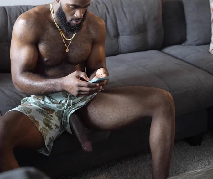 Marshall Price sitting on a brown fabric sofa while on his iPhone scrolling and his long fat dick is hanging out of his printed patterned shorts