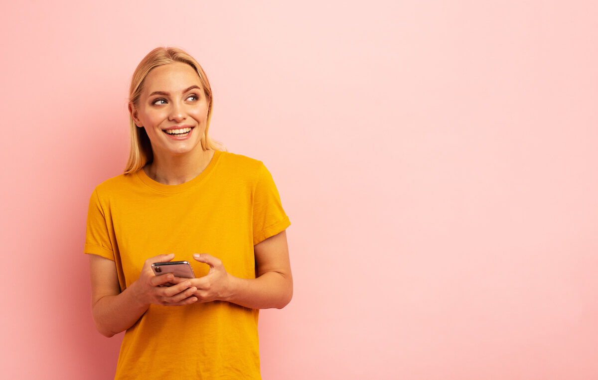 Woman in yellow shirt typing on smart phone while smiling and looking out of frame. 