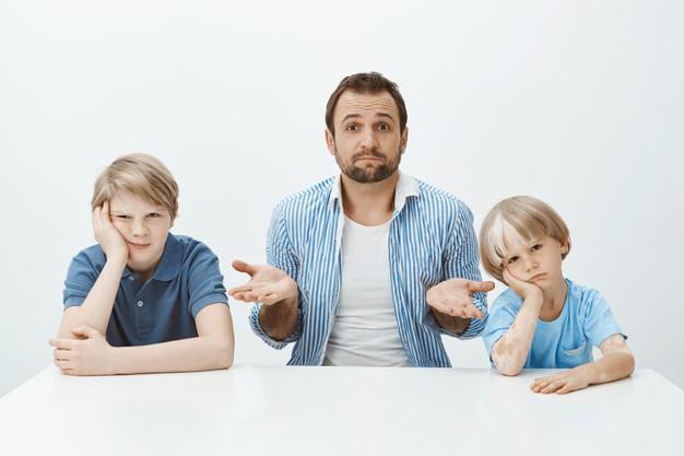 Portrait of questioned good-looking european father sitting at table with bored and upset sons, shrugging with raised palms, being clueless how to raise boys alone Free Photo