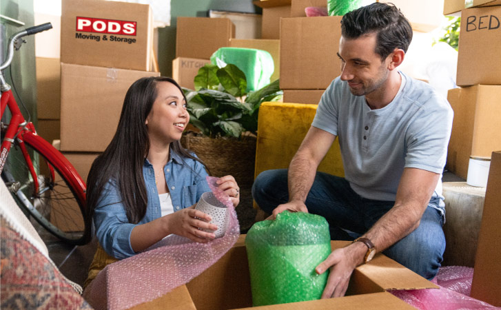 A couple surrounded by moving boxes wraps fragile vases in bubble cushioning roll before packing them up for their upcoming move into their new home.