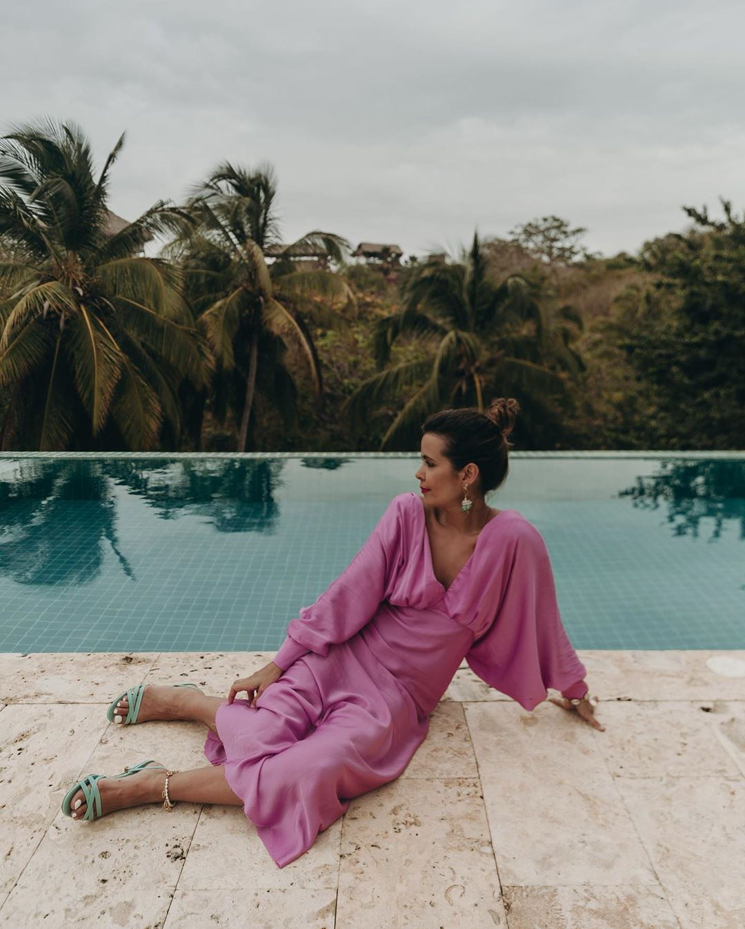 La influencer Sara Escudero posa sentada al borde de una piscina con un vestido rosa y sandalias azules, uno de los mejores look de las influencers para copiar esta temporada