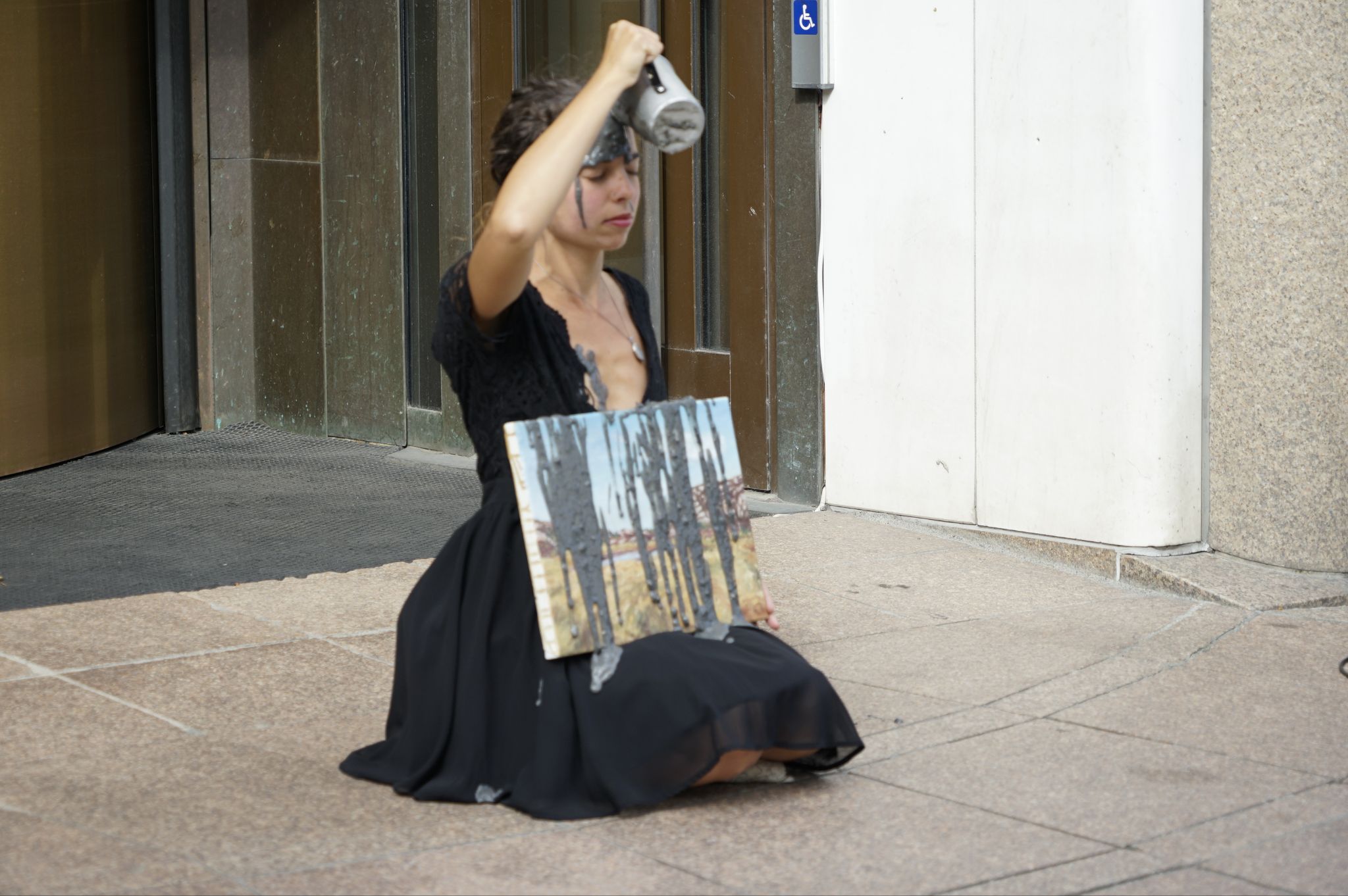 A women in a black dress kneels on the pavement holding a landscape painting and calmly pours grey liquid over herself.